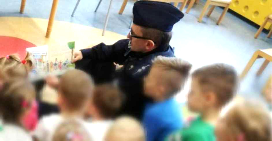 Na zdjęciu widoczny dzielnicowy w mundurze, który czyta książkę dzieciom, dzieci mają rozmyte twarze. 