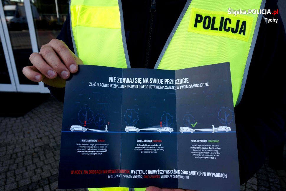 Policjant w odblaskowej kamizelce trzyma w rękach ulotkę akcji "Twoje Światła-Nasze Bezpieczeństwo". 