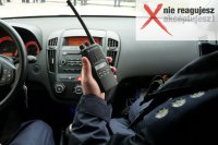 Zdjęcie przedstawia kawałek munduru policjanta siedzącego wewnątrz radiowozu, trzymającego radiostację, w prawym górnym rogu napis: &quot;Nie reagujesz-akceptujesz&quot;