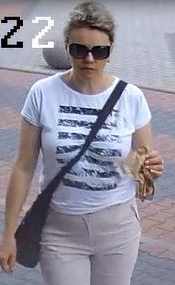Kobieta w okularach, blond włosy, biała koszulka w czarne pasy, jasne spodnie, czarna torebka przewieszona przez ramię.