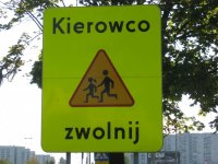 żółty znak drogowy z namalowanymi dziećmi biegnącymi przez jezdnię w tle widać zielone liście drzew