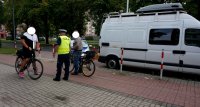 Policjant z drogówki w żółtej kamizelce stoi pomiędzy dwoma rowerzystami i rozmawia z nimi, obok zaparkowany jest radiowóz i  biały samochód.