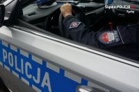 Na zdjęciu widoczny kawałek munduru z emblematem wydziału ruchu drogowego oraz radiowóz z napisem &quot;POLICJA&quot;.