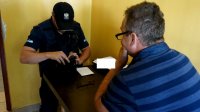 Na zdjęciu widoczny umundurowany policjant, który trzyma portfel, a na przeciwko niego, przy stoliku siedzi mężczyzna w okularach w koszulce w niebieskie paski.