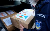 Na zdjęciu widoczna ręka policjanta w mundurze, który wkłada do bagażnika karton, na którym są rysunki dzieci.