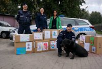 Na zdjęciu widoczni policjanci, pies służbowy oraz przedstawicielka schroniska z Tychów. Wszyscy stoją przy rozłożonych kartonach z darami dla zwierząt, w tle widoczny radiowóz.