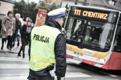 Na zdjęciu widoczny policjant, który stoi tyłem w kamizelce odblaskowej i kieruje ruchem, z naprzeciwka widoczny autobus z napisem &quot;Na cmentarz&quot; w tle widoczne inne osoby.