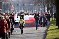 osoby uczestniczące w przemarszu ulicami miasta trzymają w pozycji poziomej dużą biało-czerwoną flagę Polski