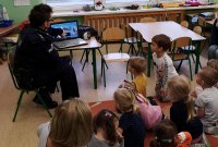 Na zdjęciu dzielnicowy pokazuje palcem na ekran komputera, gdzie wyświetlana jest prezentacja na temat bezpieczeństwa dzieciom z przedszkola, które siedzą obok i słuchają policjanta.