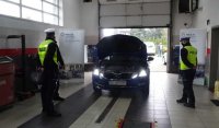 Pomiędzy policjantami na stacji diagnostycznej zaparkowany jest pojazd z włączonymi światłami.