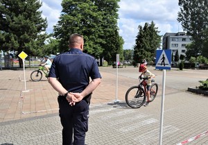 Na zdjęciu umundurowany policjant stojący tyłem. Przed nim dwoje dzieci jeżdżących na rowerach. Dzieci mają założone kaski rowerowe.