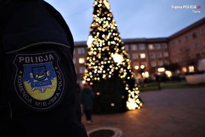 Na zdjęciu naszywka na rękawie policyjnego munduru z napisem Komenda Miejska Policji w Tychach. Z tyłu choinka ze światełkami.