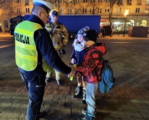 Na zdjęciu umundurowany policjant wręczający odblaski dzieciom.