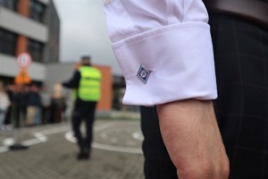 Na zdjęciu spinka na białej koszuli z logo ruchu drogowego. W tle policjant ruchu drogowego.